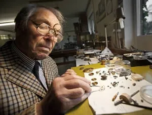 George Daniels a műhelyasztalánál, órarészletekkel a kezében és az asztalon.